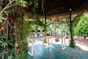 An outdoor dining area with a table set for four, surrounded by lush greenery and a thatched roof structure. - Umbrella Inn (Mahabaleshwar)