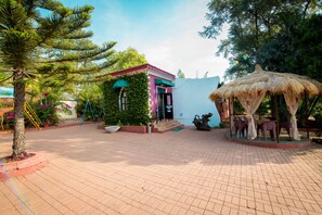 A courtyard with a small building, a thatched gazebo, and a tree. - Umbrella Inn (Mahabaleshwar)