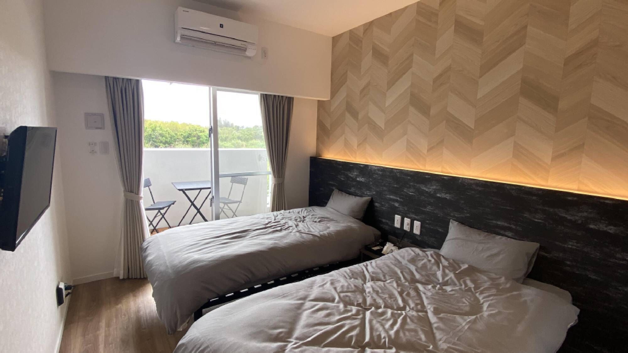 A hotel room with two beds, an air conditioning unit, a TV, and a balcony with a table and chairs.