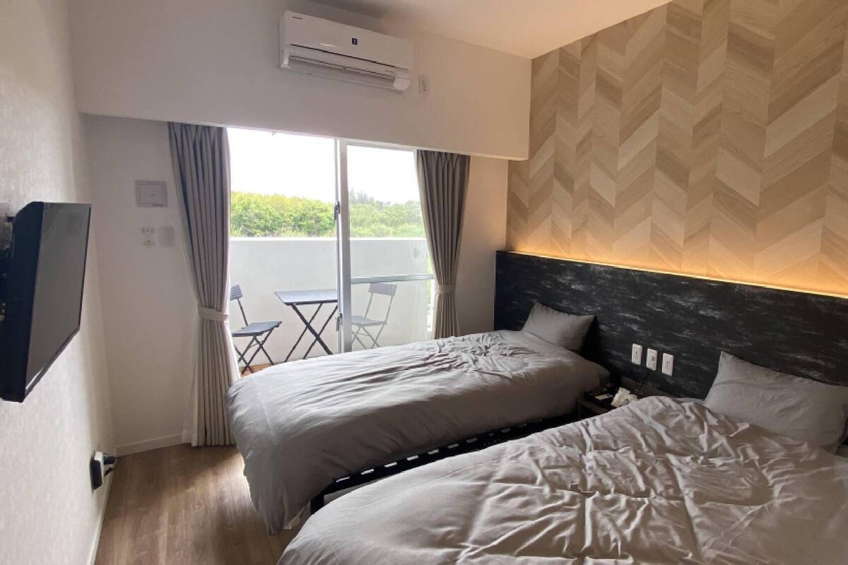 A hotel room with two beds, an air conditioning unit, a TV, and a balcony with a table and chairs.