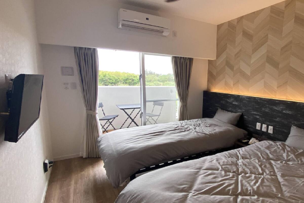A hotel room with two beds, a TV, an air conditioner, and a view of greenery through the window.