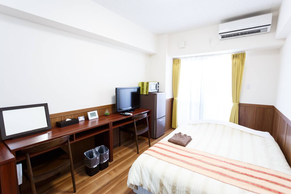 Habitació d'hotel amb llit, escriptori amb televisió, nevera i finestra amb cortines.