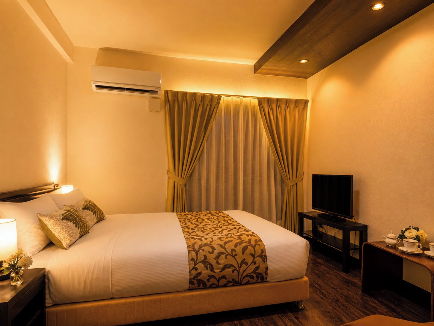 Una habitación de hotel con cama, aire acondicionado, televisión, mesita de noche con lámpara y cortinas.