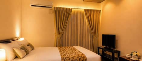 A hotel room with a bed, an air conditioning unit, a television, a bedside table with a lamp, and curtains.