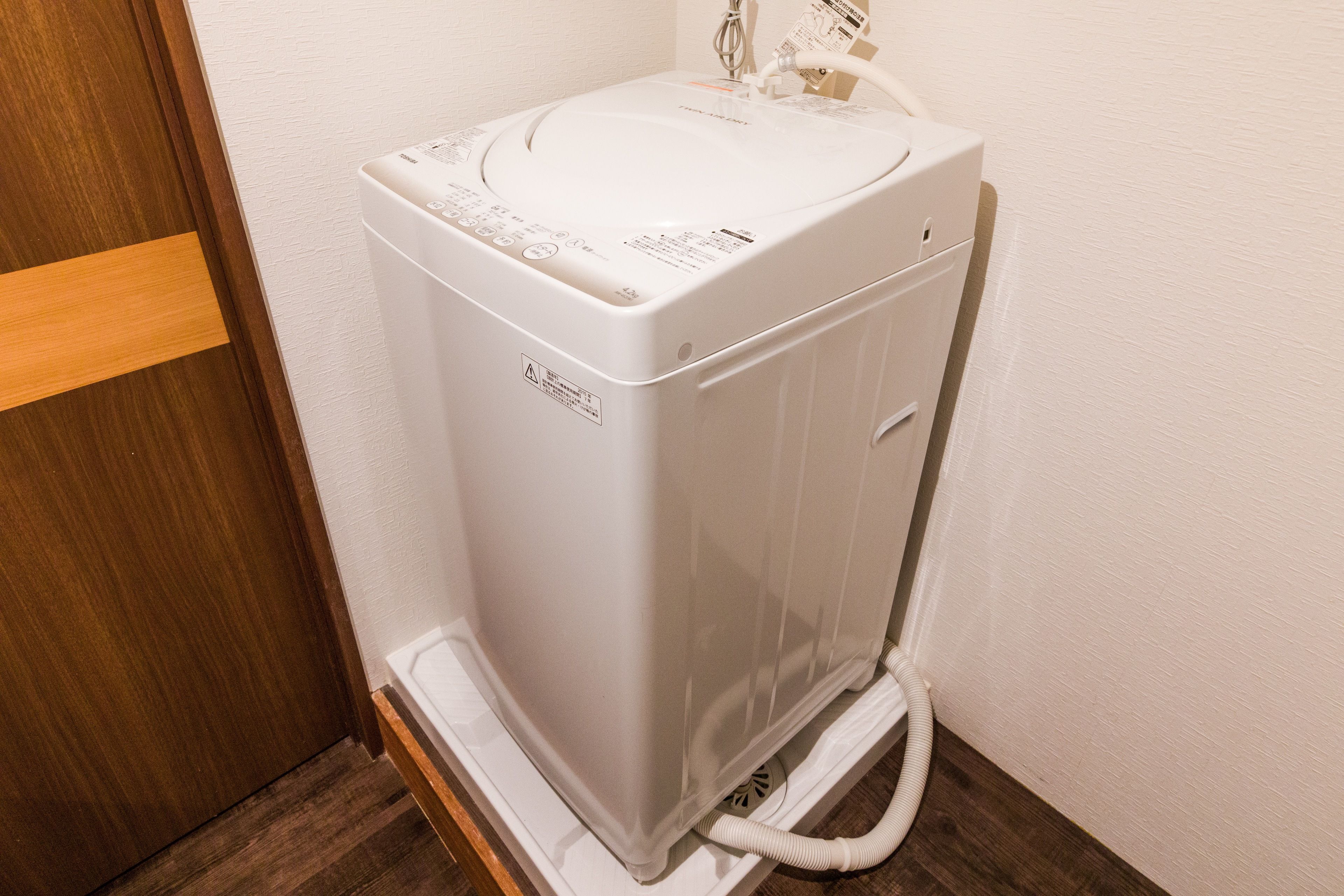A compact, white, wall-mounted washing machine in a corner with a wooden door frame.