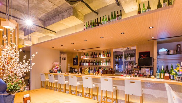 A bar area with multiple seating options, a variety of bottles on shelves, and a television screen. - Mr.KINJO in MATSUYAMA (Naha)