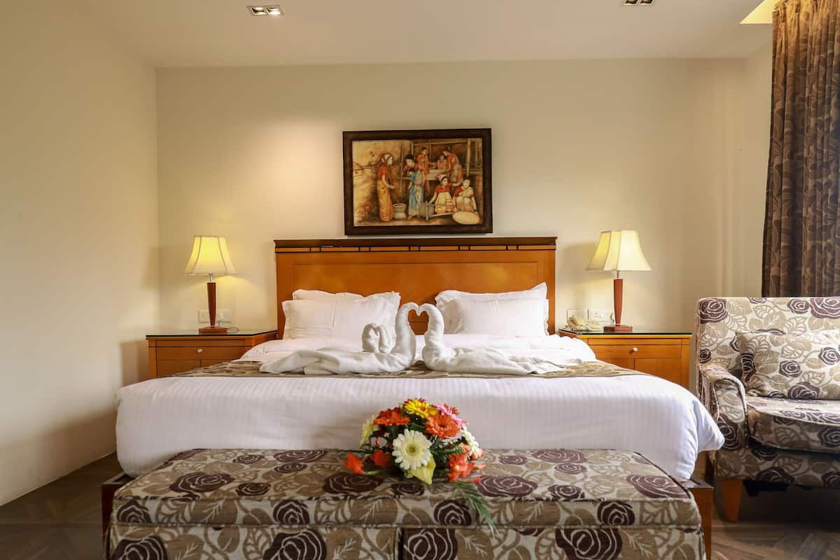 A hotel room with a bed, two bedside lamps, a painting above the bed, a floral arrangement on the bed, and a patterned ottoman.