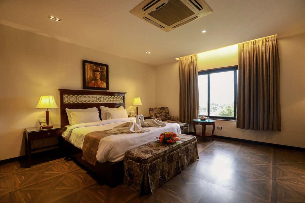A hotel room with a bed, two bedside lamps, a nightstand, a small table, a window with curtains, and a wall-mounted air conditioner.