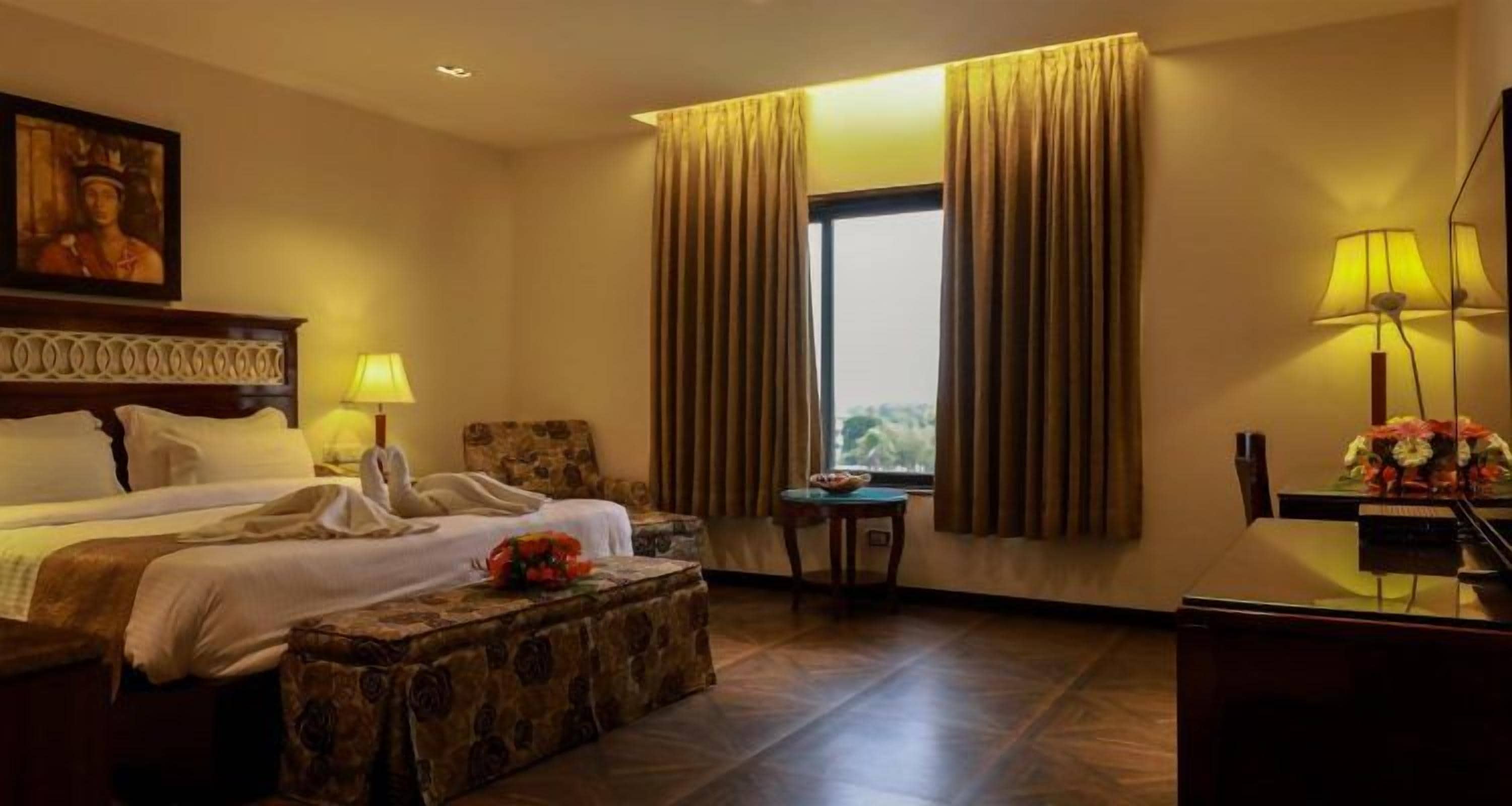A hotel room with a bed, a desk, a chair, a window with curtains, and a painting on the wall.