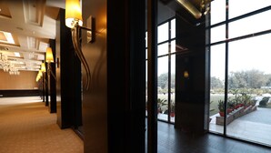 A hotel corridor with a carpeted floor, wall-mounted lamps, and large windows offering a view of the outdoors.