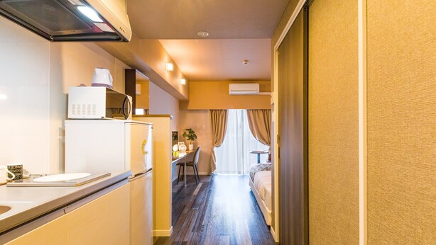 A narrow hallway with a kitchenette on the left, a dining area with a table and chairs, and a bed with curtains on the right. - Mr.KINJO in Asahibashi station square (Naha)