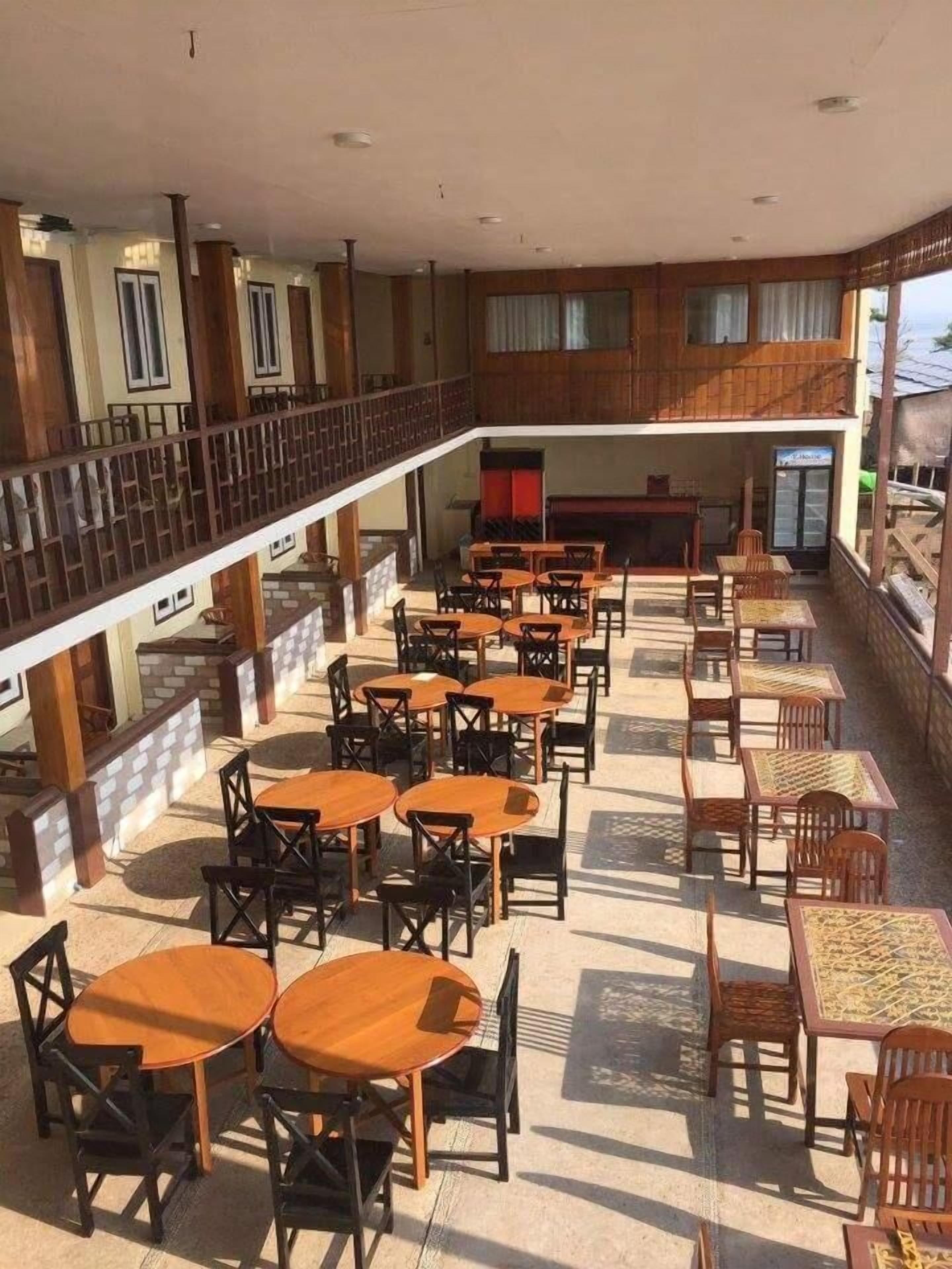 A spacious indoor area with multiple wooden tables and chairs arranged in rows, a balcony with a railing, and large windows allowing natural light.