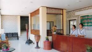 A reception area with a wooden reception desk, two receptionists, a potted plant, a traditional drum, and a brick wall.