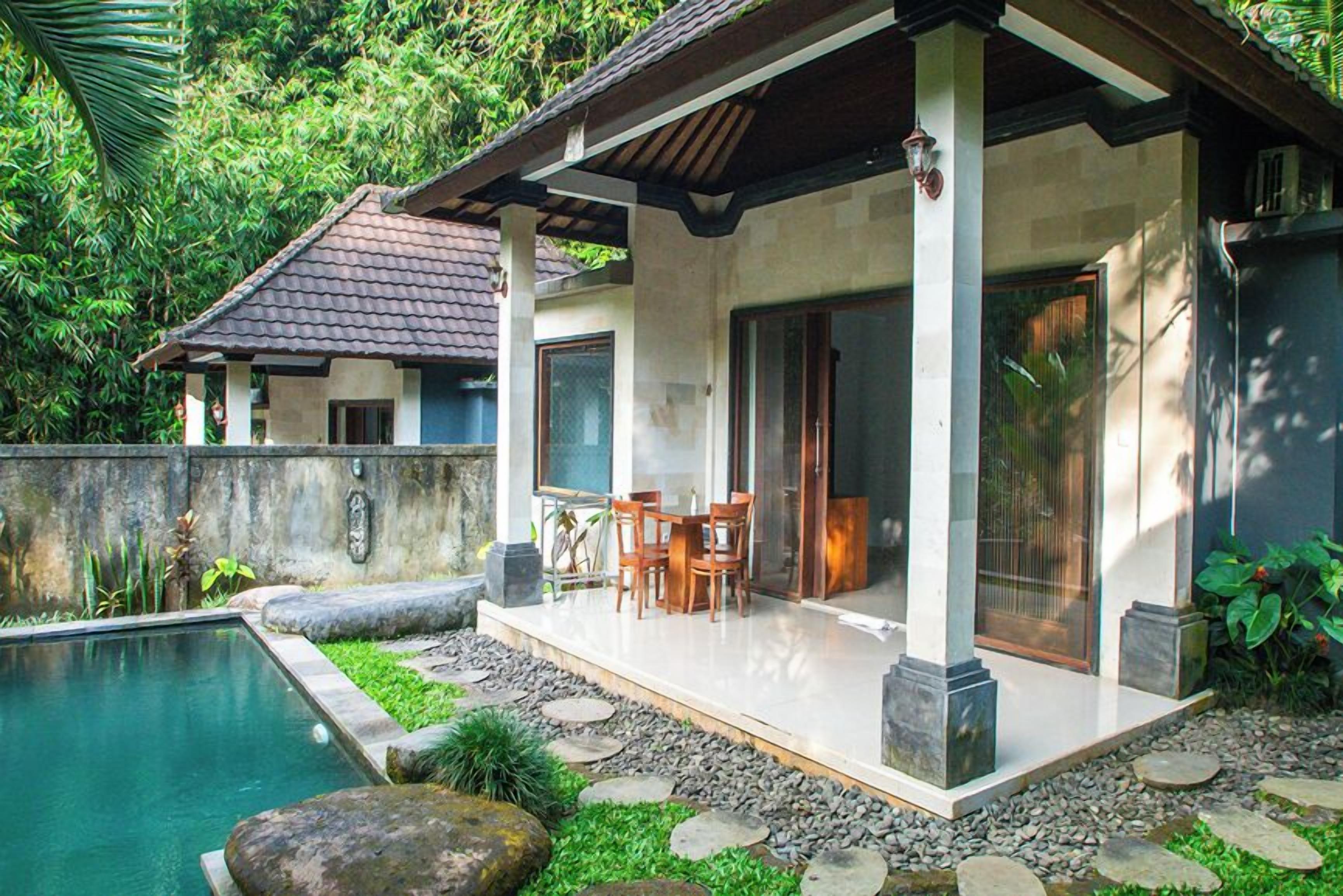 A poolside area with a covered patio, stone pathway, and a view of a house with a tiled roof.