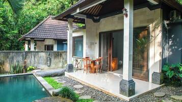 A poolside area with a covered patio, stone pathway, and a view of a house with a tiled roof.