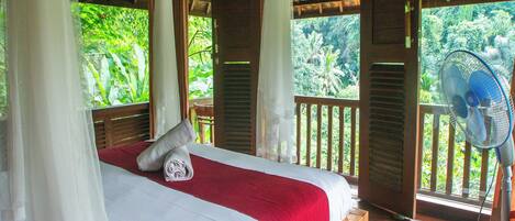 A bedroom with a wooden bed, white and red bedding, a ceiling fan, and a view of lush greenery outside.