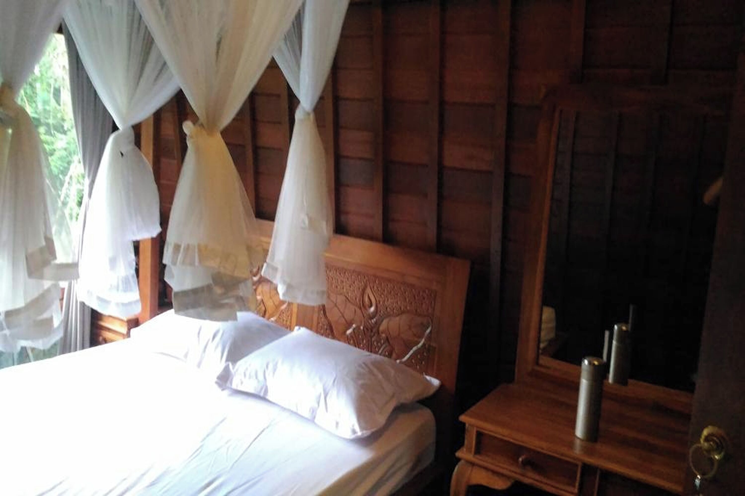 Een houten bed met wit beddegoed en een hemelbed, een houten nachtkastje met een spiegel en een kaars, en een houten hoofdbord met een gegraveerd ontwerp.