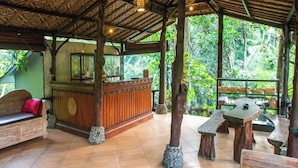 A wooden reception desk with a mirror, a bench, and a table in a covered outdoor area with a tiled floor. - Bali Jungle Resort (Sebatu)