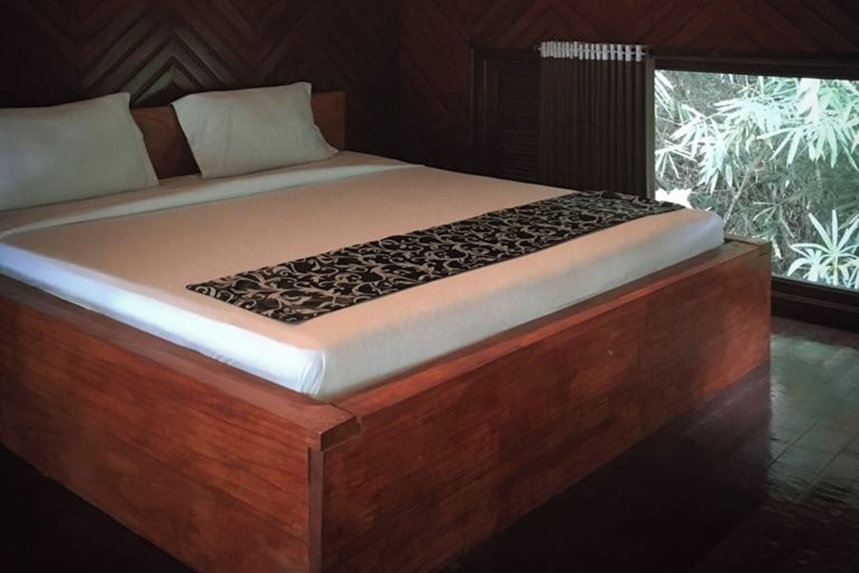 A large bed with a patterned runner, wooden headboard, and a window with a view of foliage.