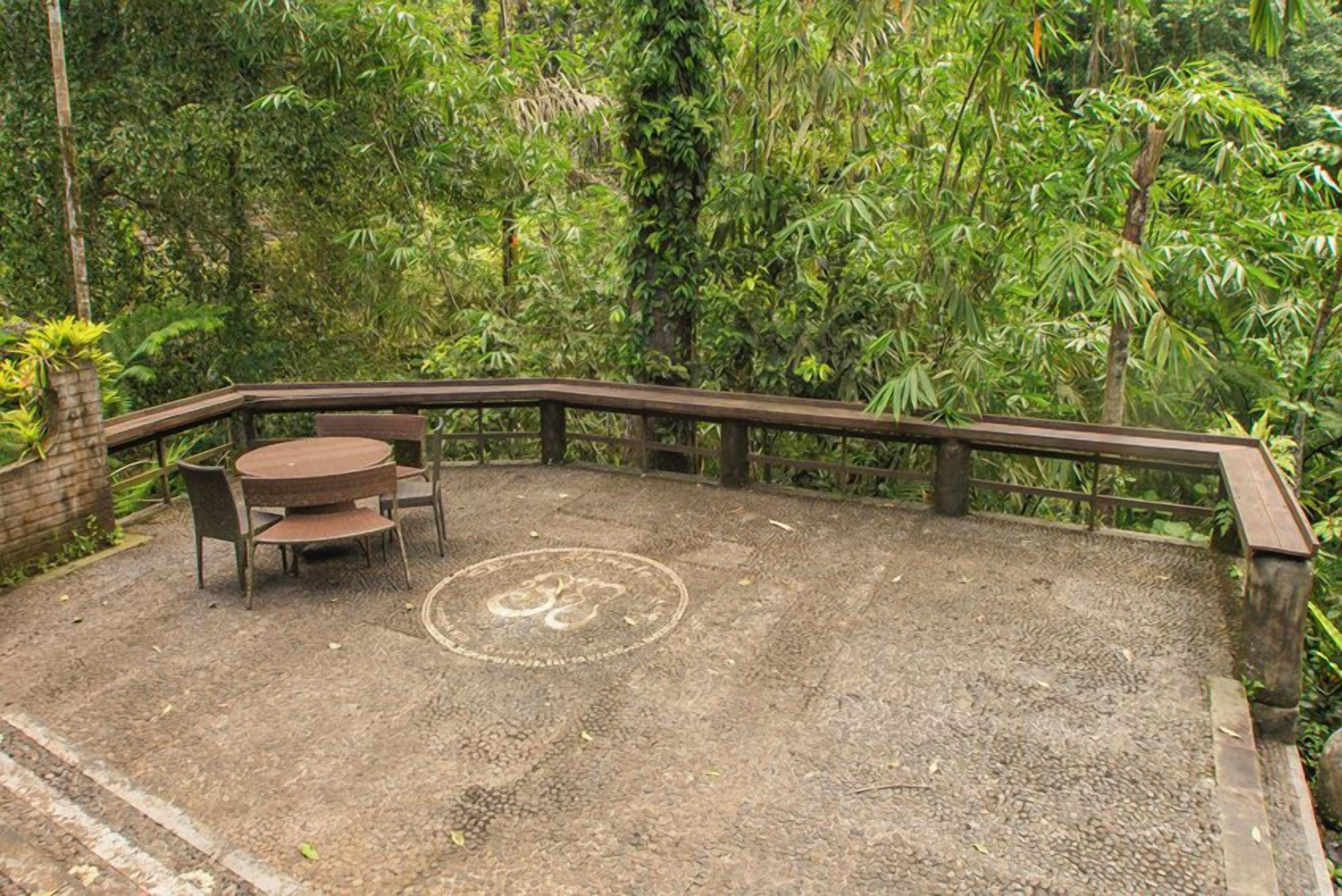 An outdoor seating area with a round table and chairs, surrounded by a wooden fence and dense foliage.