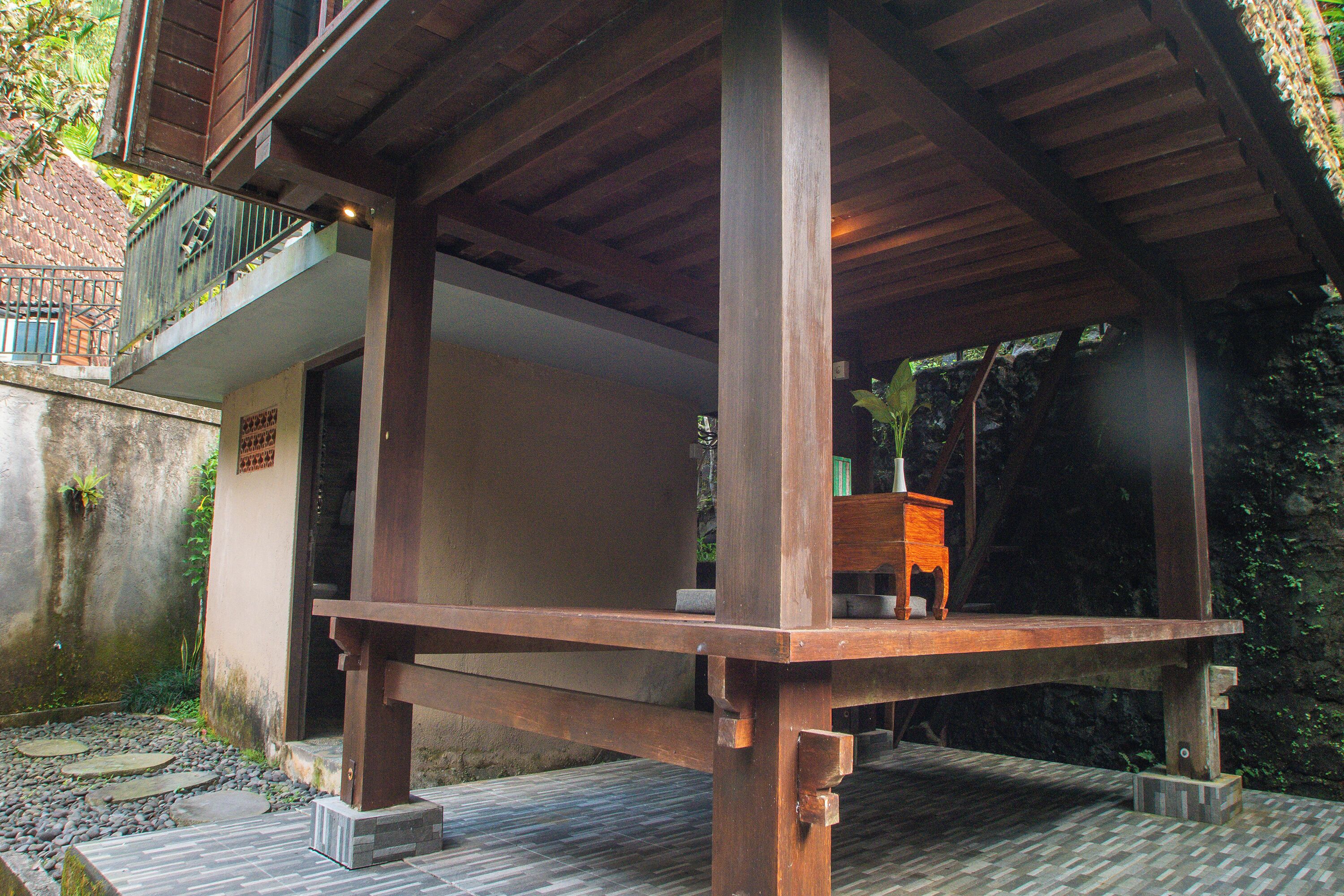 A wooden structure with a bench and a small table, surrounded by a stone pathway and greenery.