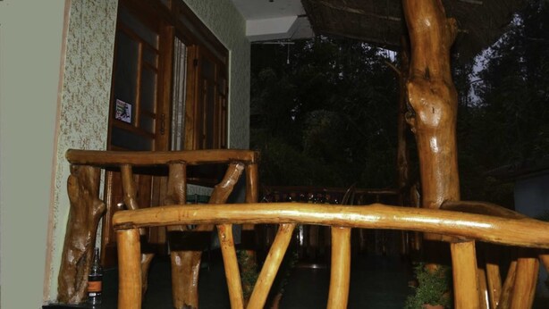 Una silla de madera rústica con acabado natural, ubicada en un espacio al aire libre con techo de paja y vistas a árboles en el fondo.