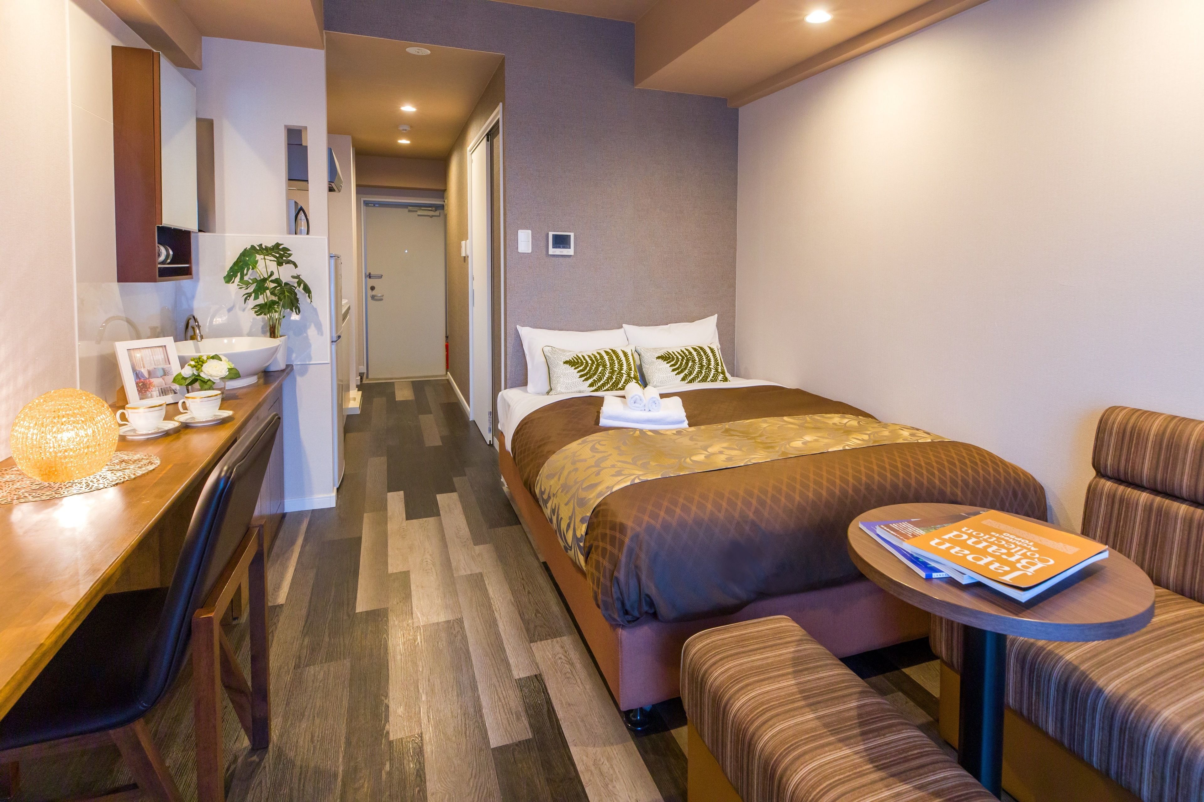A hotel room with a bed, a bedside table with a book, a desk with a lamp, and a chair.