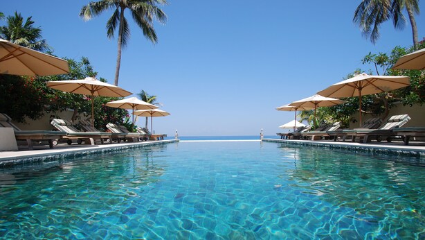 Ein klares Schwimmbecken mit Liegestühlen und Sonnenschirmen, Palmen und Blick auf den Ozean.