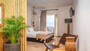 A hotel room with a bed, a potted plant, a wooden chair, a table, and a view of the sea through an open door. - Ariadne Hotel (Naxos)