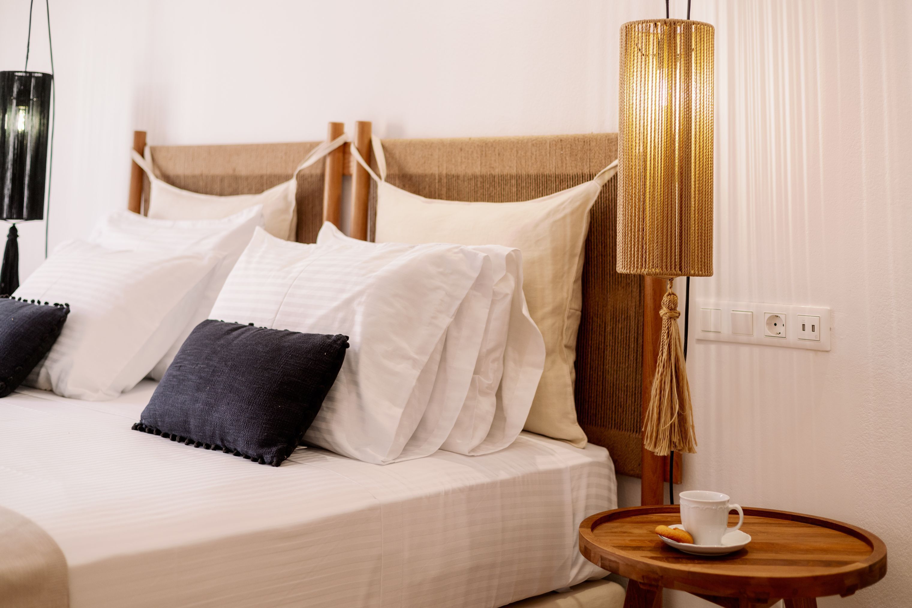 A neatly made bed with white and dark pillows, a round wooden table with a cup and plate, and a tall, textured lamp beside the bed.