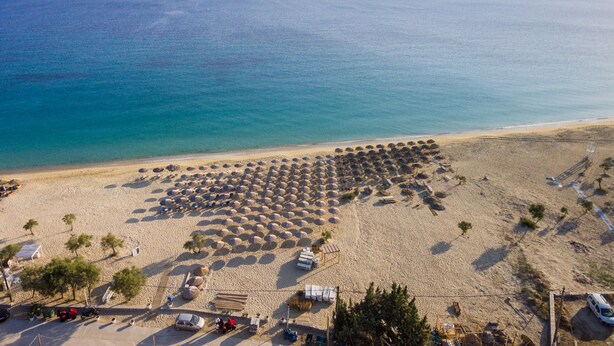 An aerial view of a beach with rows of umbrellas and beach chairs, a parking area, and a clear view of the sea and distant mountains. - Ariadne Hotel (Naxos)