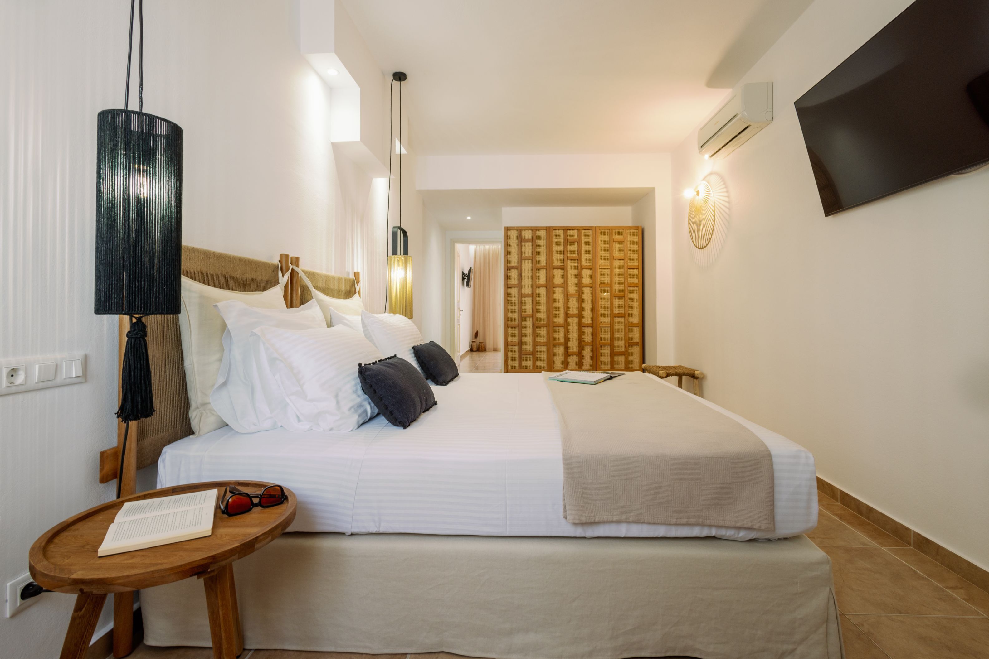A hotel room with a bed, a bedside table with glasses and a book, a lamp, a TV, and a wall-mounted air conditioner.