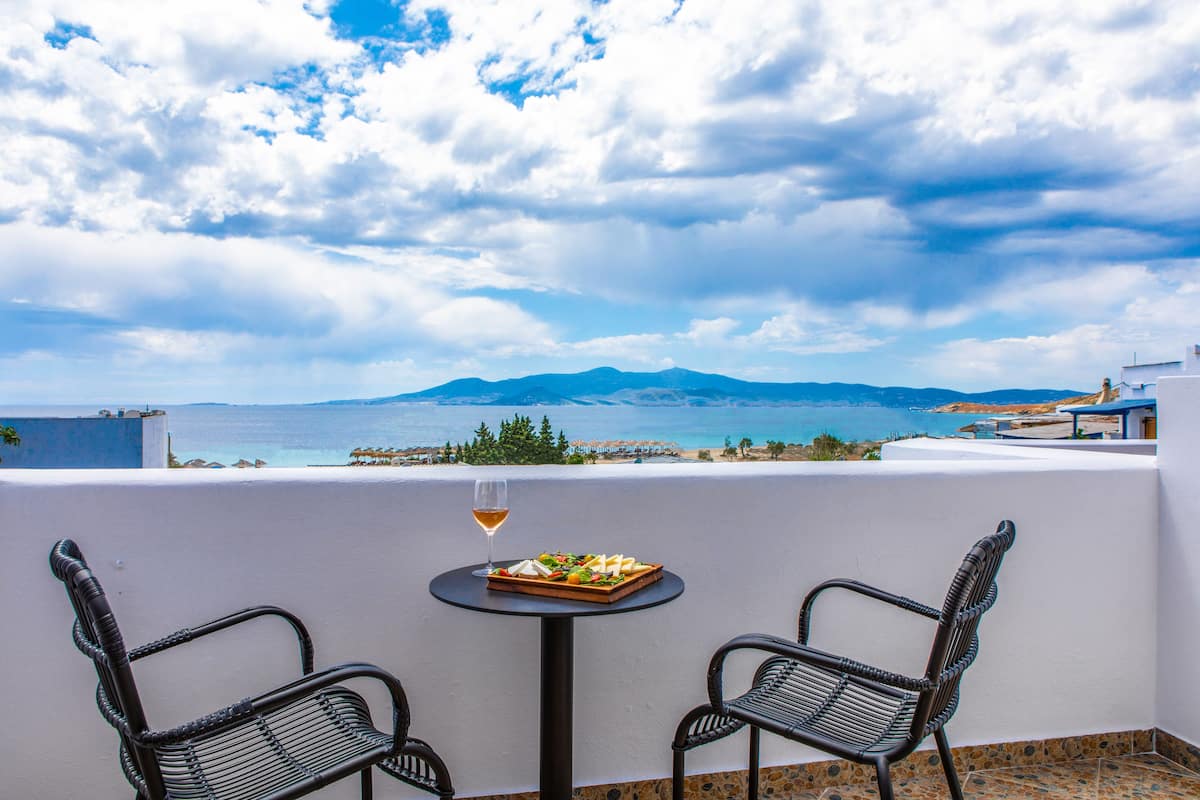 Un balcón con dos sillas de mimbre, una pequeña mesa redonda con una copa de vino y un plato de comida, con vistas a un paisaje costero con montañas al fondo.