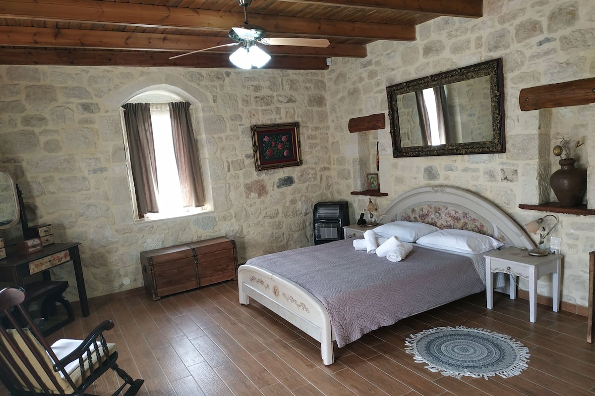 Un dormitorio con pared de piedra, suelo de madera, una cama con cabecera, una mesita de noche, una silla, un aparador, un espejo y un ventilador de techo.