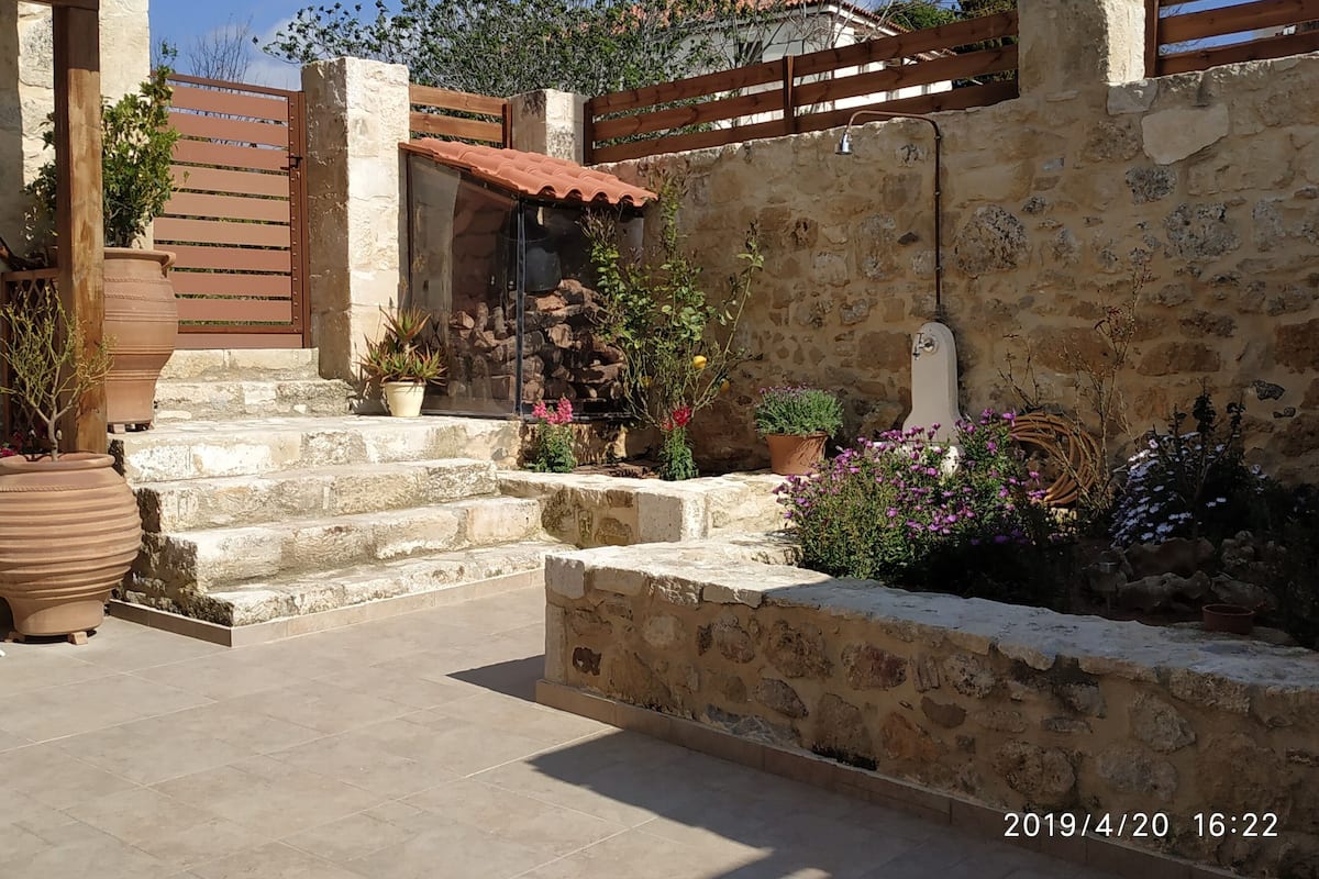 Un patio con muros de piedra y escalones que conducen a un portón de madera, plantas en macetas y una fuente.
