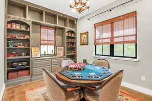 Une pièce avec une table ronde préparée pour des jeux de cartes, des étagères remplies d’objets de collection et une fenêtre avec des stores.