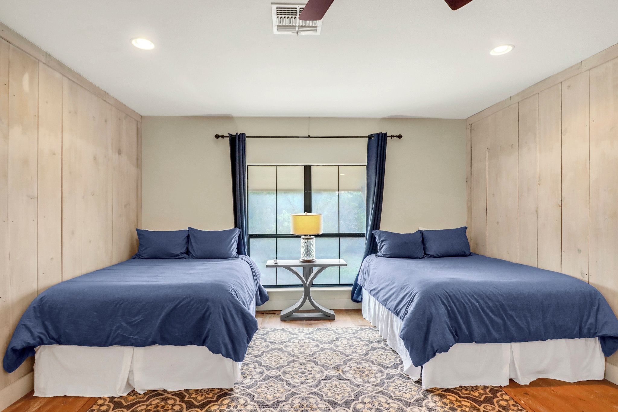 A room with two beds, a small table with a lamp, a patterned rug, and a window with curtains.
