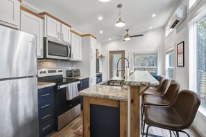 Cocina moderna con gabinetes blancos, electrodomésticos de acero inoxidable, encimera de mármol y una zona de bar con sillas marrones.