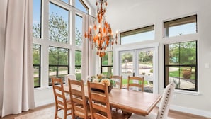 A dining room with a large wooden table, six chairs, a chandelier, and large windows offering a view of the outdoors. - 4BR Canyon Lake Retreat with New Private Pool & Outdoor Living (Canyon Lake)