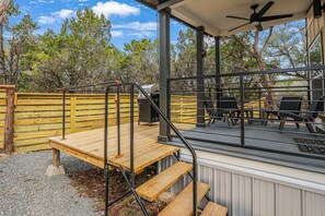 Una terraza techada al aire libre con escalones de madera y un ventilador de techo, rodeada de árboles.