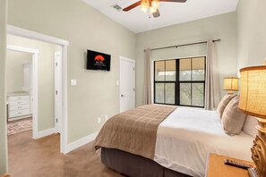 Une chambre à coucher avec un lit, une télévision, une fenêtre avec des rideaux, un ventilateur de plafond et une lampe.