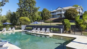 A swimming pool with lounge chairs and umbrellas, surrounded by trees and a building with multiple balconies.