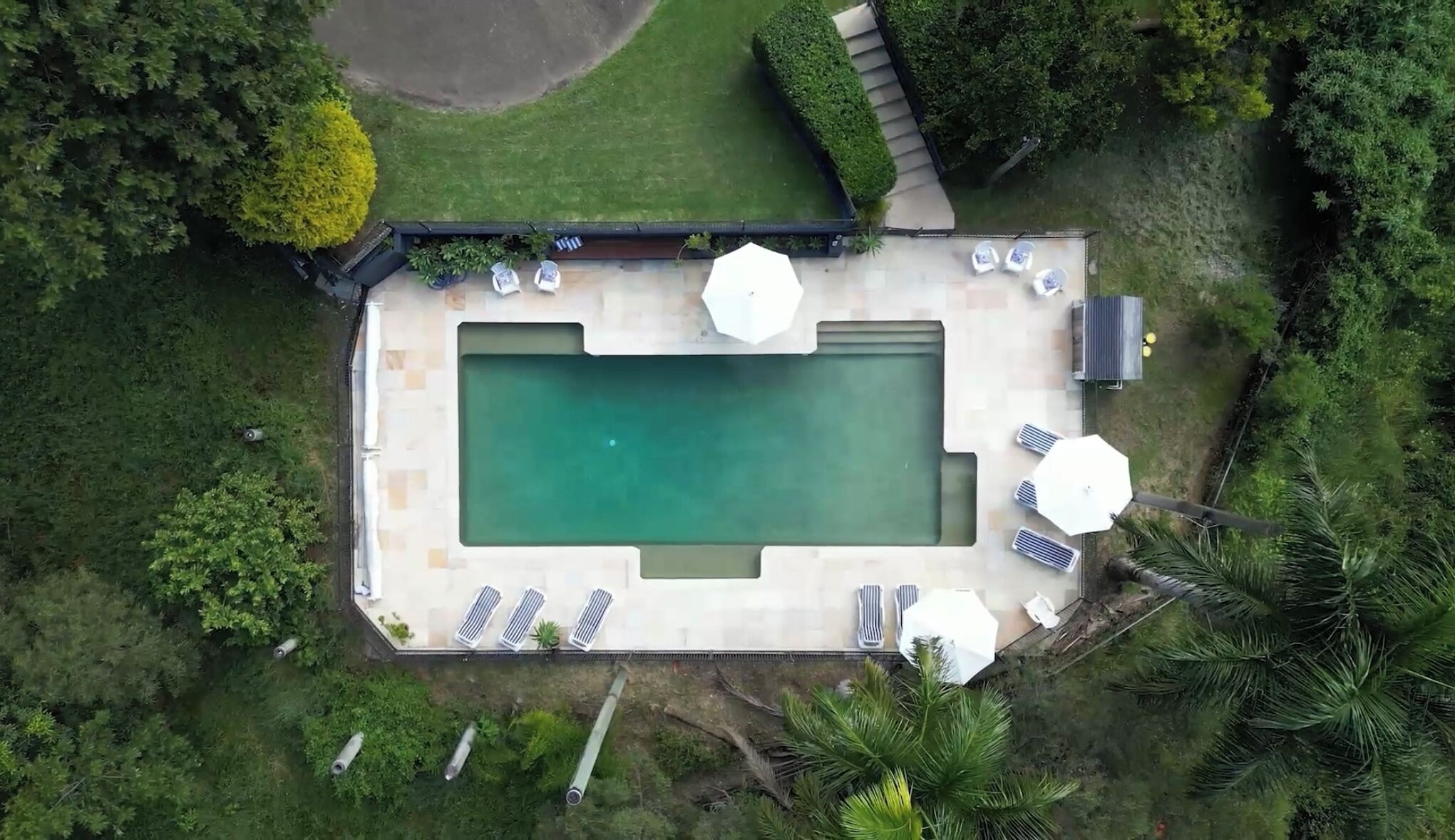 An aerial view of a rectangular swimming pool with a tiled deck, surrounded by lounge chairs and umbrellas, set in a lush green landscape.
