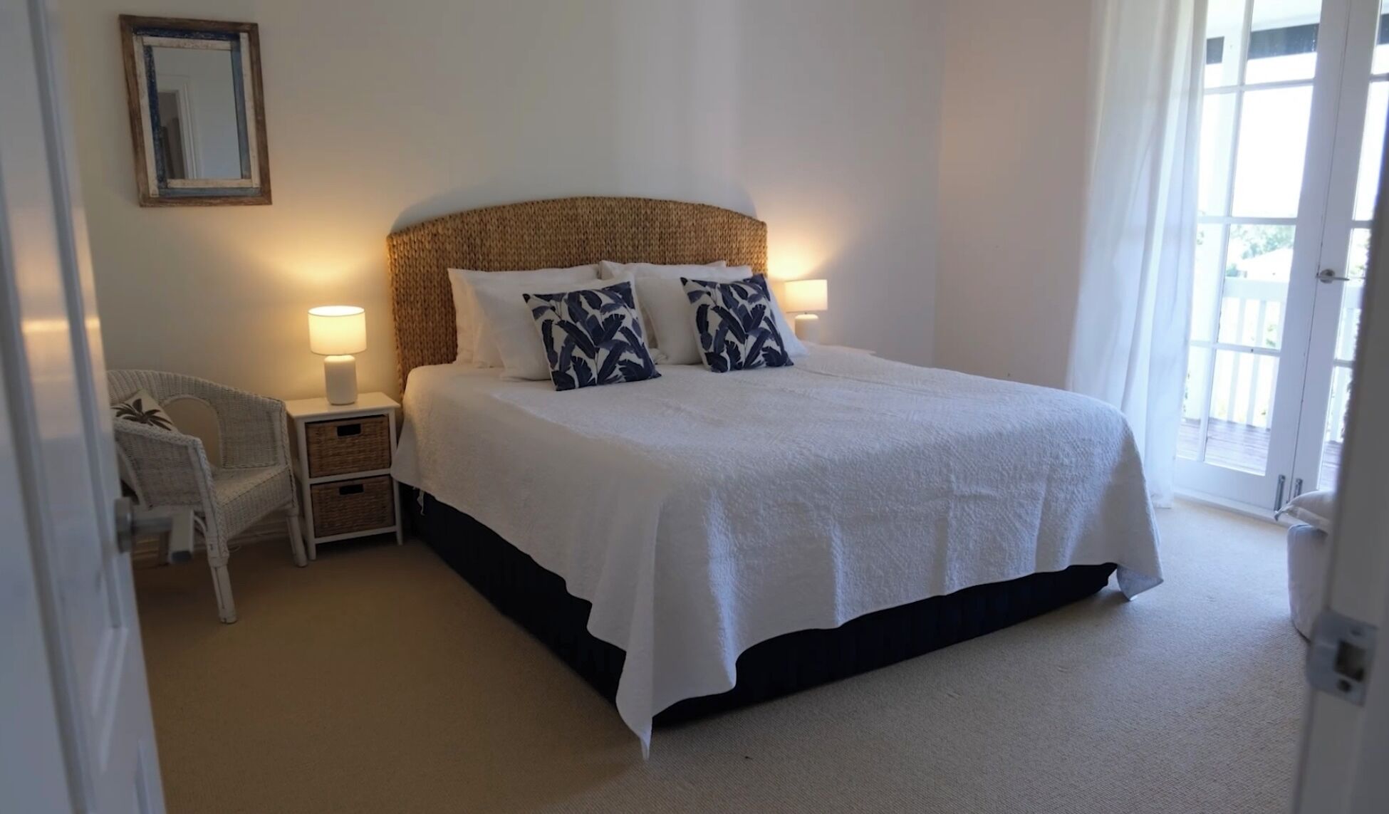 A neatly made bed with white bedding and two decorative pillows, a bedside lamp, and a wicker chair in a well-lit room with a large window.