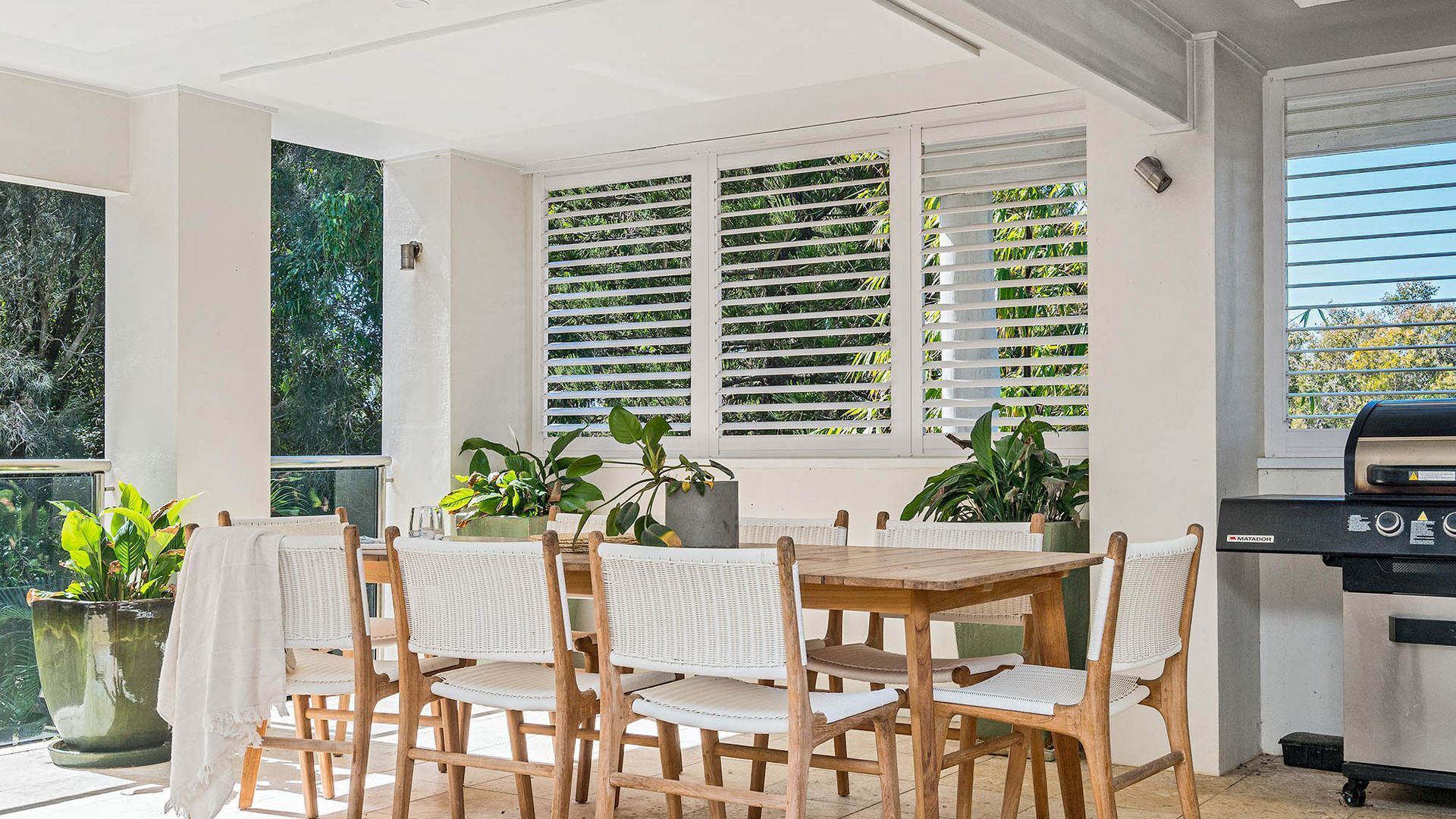 A covered patio area with a wooden table and chairs, potted plants, and a grill.