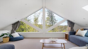 Une pièce moderne et bien éclairée, avec un plafond en pente, de grandes fenêtres donnant sur la verdure, un canapé bleu, une petite table et une plante en pot.