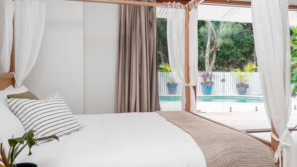 A bedroom with a large bed, white curtains, and a view of a swimming pool and greenery outside.
