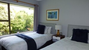 A hotel room with two beds, a window with a view of trees, a framed picture on the wall, and a nightstand with a lamp. - Tasteful spacious private garden quest house (Katikati)