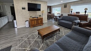 A spacious living room with a wooden coffee table, grey sofas, and a flat-screen TV mounted on the wall. - Near town & Golf, Sleeps 15, 3,300sqft, 1.9 Acres, 6BR w/4Kings, Game Room, HVAC (Williams)