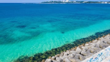A coastal view with clear turquoise waters, a rocky breakwater, and a distant cityscape.